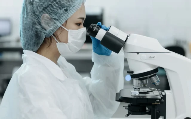 Cercetătoare cu echipament de protecție care folosește un microscop într-un laborator.