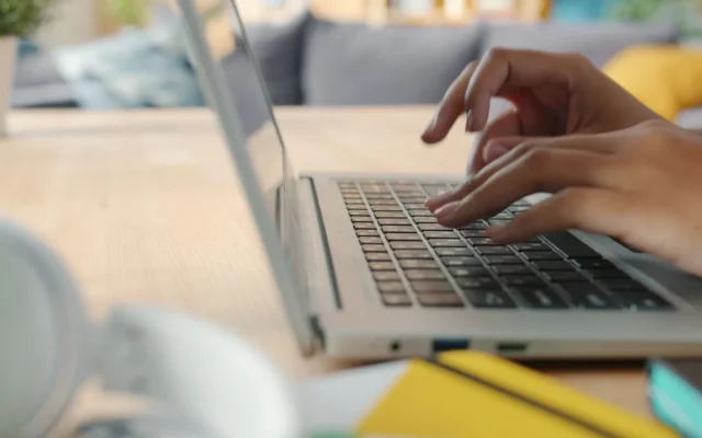 Mâini tastând pe tastatura unui laptop, cu fundal neclar