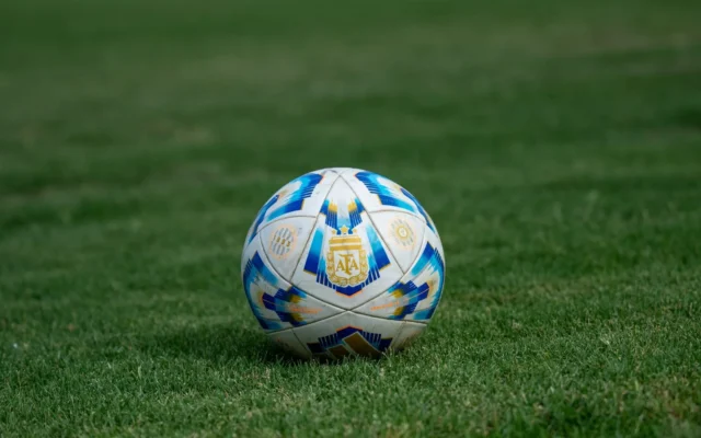 Minge albă de fotbal, cu model albastru și auriu și emblema AFA, pe gazon