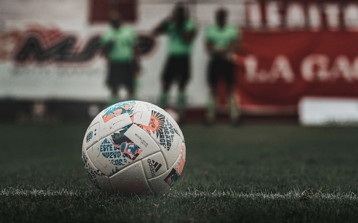 Minge albă de fotbal pe gazon, cu persoane în echipament verde în fundal