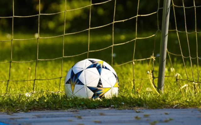 Minge de fotbal albă, cu modele albastre și galbene, pe gazon lângă plasa porții