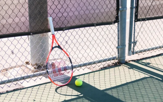 Rachetă și minge de tenis pe teren, lângă un gard metalic
